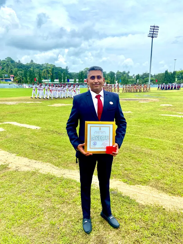 Hardik in a dark suit holding the framed Lieutenant Governor's Commendation and medal case on the Independence Day parade ground