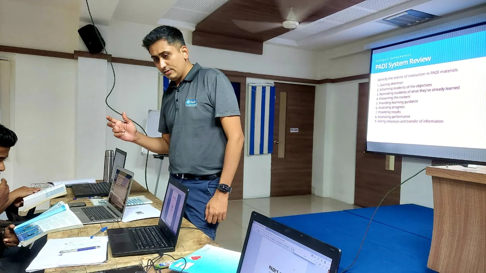 Hardik teaching an Instructor Development Course classroom session, a PADI System Review slide on screen and candidates at laptops