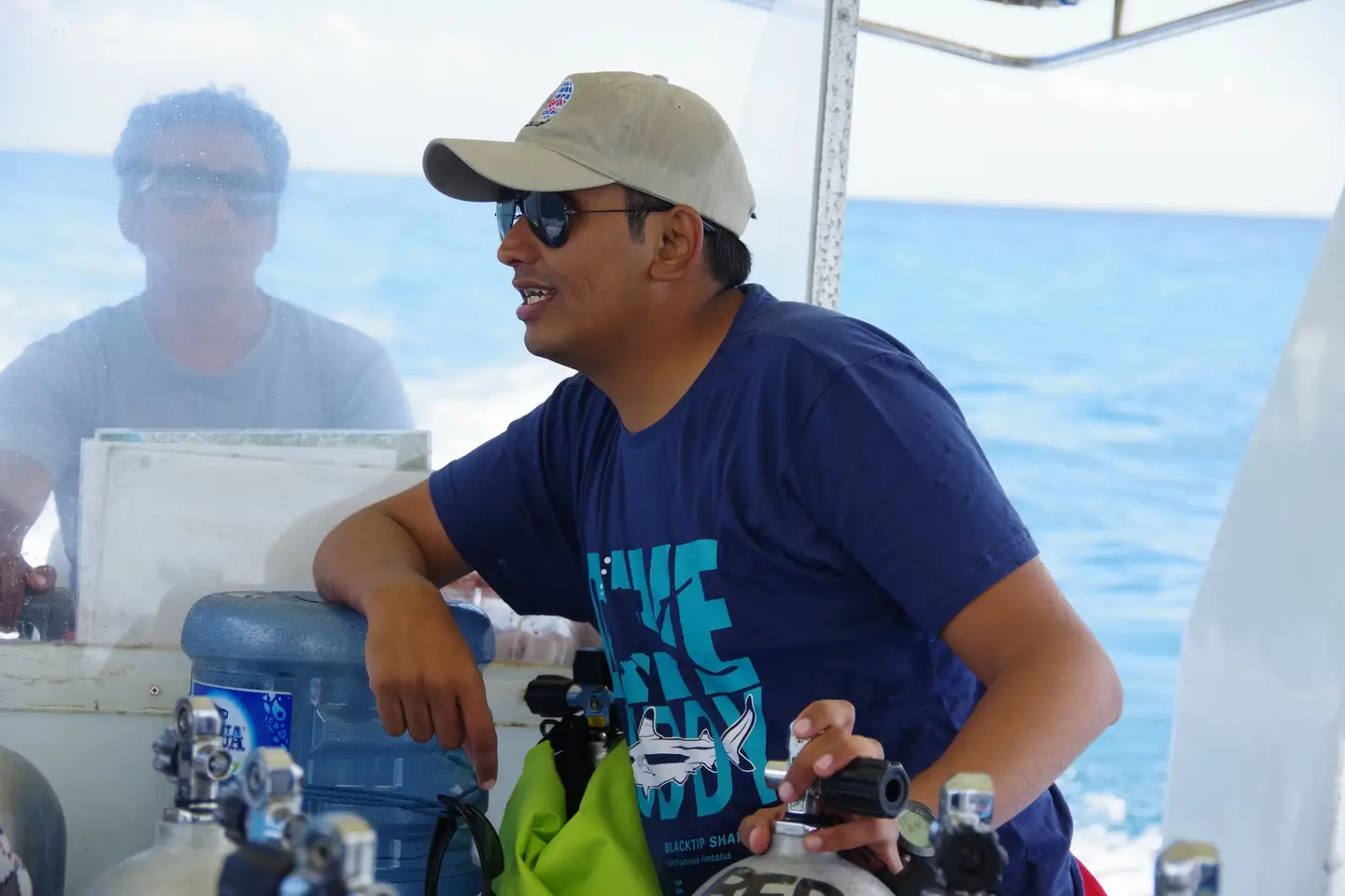 Hardik on a dive boat among cylinders and regulators, checking equipment with the sea behind